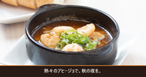 黒い小鍋の海老のアヒージョとバゲット、「熱々のアヒージョで、秋の夜を。」の文字