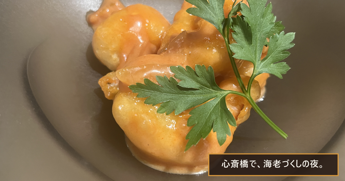 深皿に盛られた、オレンジ色のソースをまとった海老の一皿とイタリアンパセリ。「心斎橋で、海老づくしの夜。」の文字