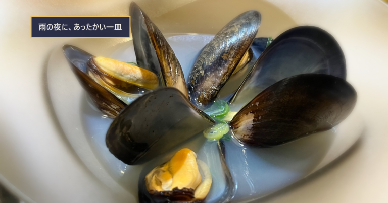 心斎橋「貝と海老そして蟹」の、白い深皿に貝が並んだあたたかいスープ仕立ての一皿。「雨の夜に、あったかい一皿」の文字